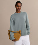 A woman in a Cashmere Oversized Crewneck in light blue and white pants stands against a beige background, confidently holding a mustard yellow suede handbag under her arm.