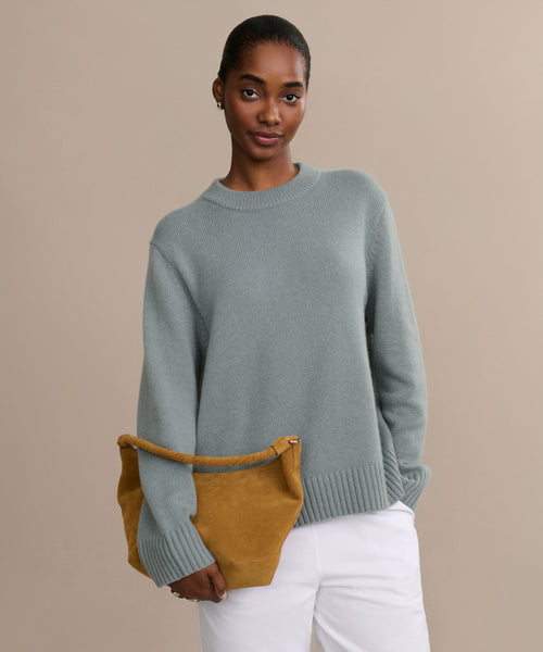 A woman in a Cashmere Oversized Crewneck in light blue and white pants stands against a beige background, confidently holding a mustard yellow suede handbag under her arm.