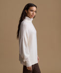 A woman wearing a cream-colored ribbed turtleneck sweater, shown in profile against a solid tan studio background.