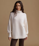 A woman modeling an oversized cream-colored knit turtleneck sweater against a solid tan studio background.