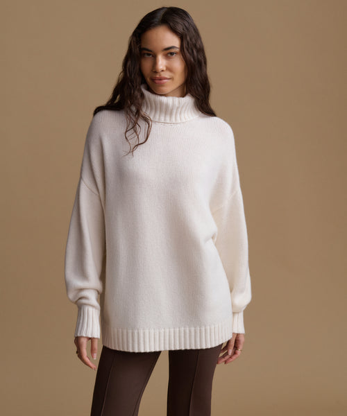 A woman modeling an oversized cream-colored knit turtleneck sweater against a solid tan studio background.