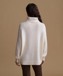 A woman viewed from the back wearing an oversized cream knit turtleneck sweater and dark brown pants, standing against a plain warm beige studio background.