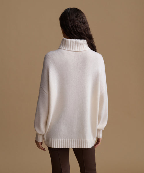 A woman viewed from the back wearing an oversized cream knit turtleneck sweater and dark brown pants, standing against a plain warm beige studio background.