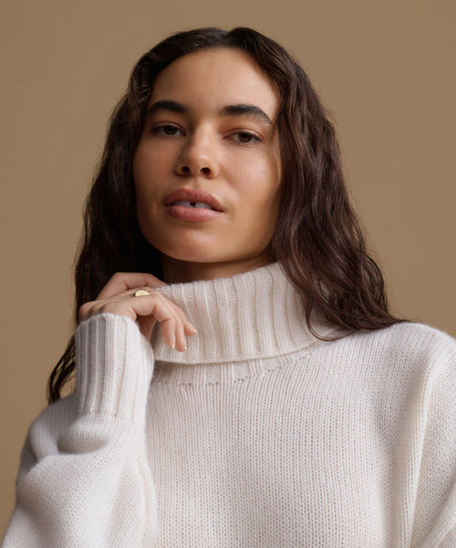 A woman wearing a cream-colored ribbed turtleneck sweater, posed against a solid warm beige background.