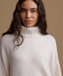 A woman wearing a cream-colored ribbed turtleneck sweater, photographed against a solid warm beige background.