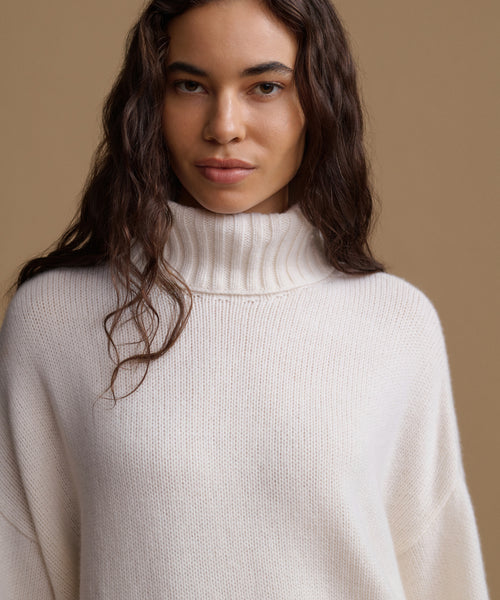 A woman wearing a cream-colored ribbed turtleneck sweater, photographed against a solid warm beige background.