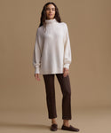 A woman modeling an oversized cream knit turtleneck sweater with dark brown cropped trousers and dark brown loafers, standing against a solid beige studio background.