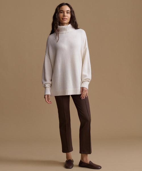 A woman modeling an oversized cream knit turtleneck sweater with dark brown cropped trousers and dark brown loafers, standing against a solid beige studio background.