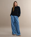 A woman with long hair stands against a beige background, wearing the Denim Newport Trouser, a navy striped sweater, and brown sandals—an effortless spring and summer look.