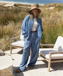 A woman stands on a patio by the beach in a wide-brimmed hat, Parker Oversized Shirt over a white top, Denim Newport Trouser, sandals, and sunglasses. Two empty chairs are beside her with grass and sand nearby.