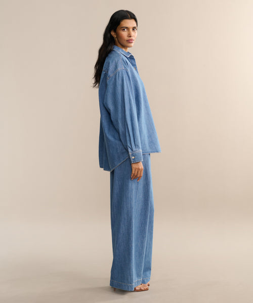 A woman with long dark hair stands barefoot in profile, wearing a Parker Oversized Shirt and Denim Newport Trouser against a plain beige background—an effortless look for spring and summer.