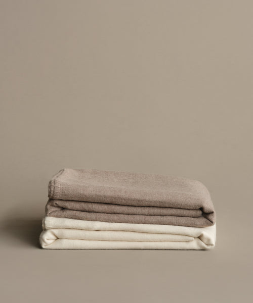Two neatly folded blankets, including the luxurious Chateau Alpaca Coverlet in ivory and a beige throw, are stacked atop each other against a plain light brown background.