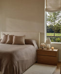 A minimalist bedroom with a beige bed topped by the Chateau Alpaca Coverlet, three matching pillows, a wooden nightstand with a lamp, flower vase, and candle, beside a window overlooking green trees and grass.