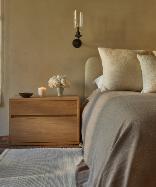 A cozy bedroom features a wooden nightstand with a candle, bowl, and white flower vase next to a bed topped with cream pillows and the Chateau Alpaca Coverlet, beneath a black wall sconce on a neutral wall.