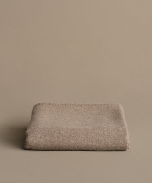 The Chateau Alpaca Coverlet, in beige, is neatly folded and placed on a matching surface with a minimalistic background.