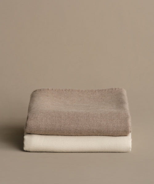 Two neatly folded Chateau Alpaca Coverlets are stacked on a plain beige background. The top is soft light brown baby alpaca, and the bottom is a cream-colored minimalist coverlet.