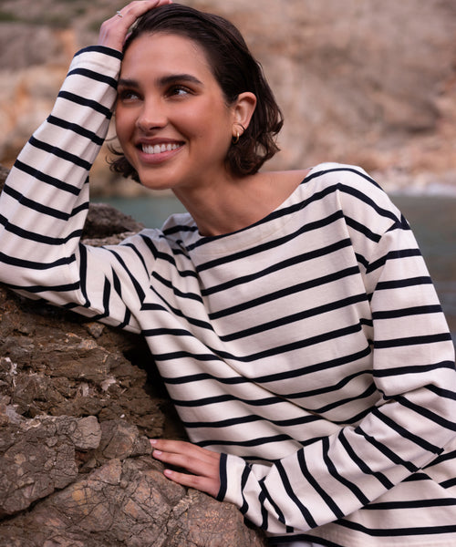 A woman smiles while leaning against a rock outdoors, wearing the Claude Long-Sleeve Tee in black and white nautical stripes. The 100% cotton top stands out against the rocky terrain and blurred natural background.