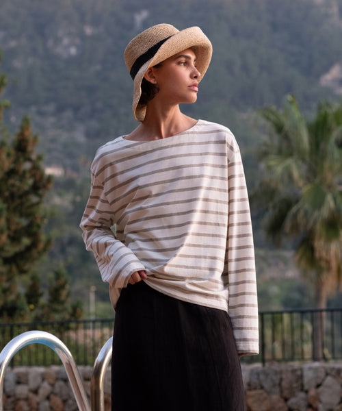A person by a pool, with trees and mountains behind them, wears a wide-brimmed straw hat, a Claude Long-Sleeve Tee in nautical stripes made of 100% cotton, and a dark skirt.