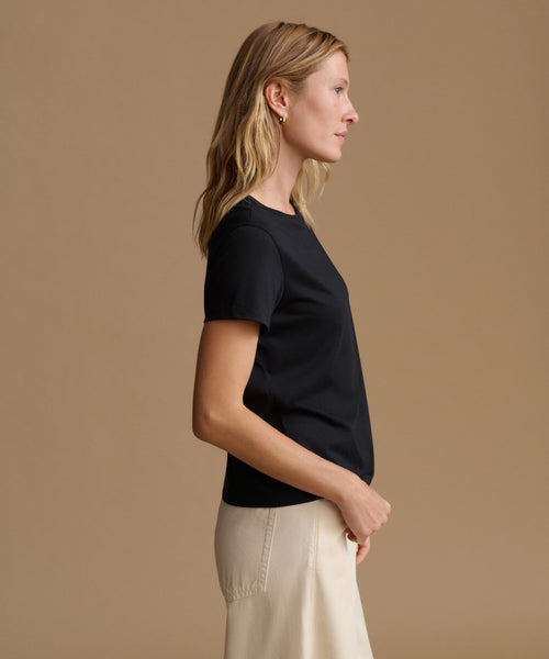 A woman in side profile wearing a black short-sleeve crewneck top and cream-colored trousers, set against a solid warm beige studio background.