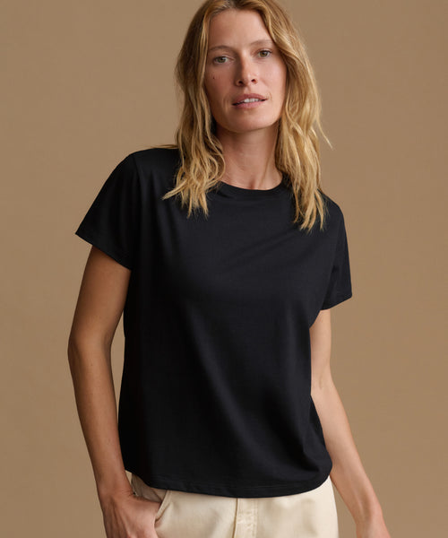 A woman wearing a black short-sleeve crewneck t-shirt and light beige pants, standing against a solid warm beige studio background.