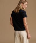 A woman shown from the back wearing a black short-sleeve crewneck t-shirt and cream-colored trousers, set against a solid warm beige studio background.