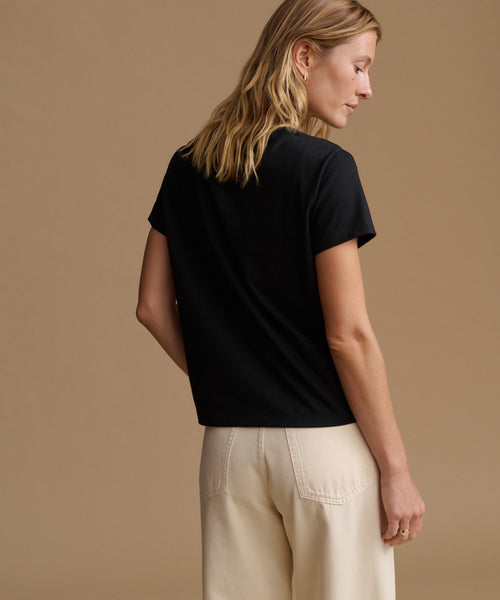 A woman shown from the back wearing a black short-sleeve crewneck t-shirt and cream-colored trousers, set against a solid warm beige studio background.