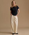 A woman stands against a plain beige studio background wearing a black short-sleeve crew-neck T-shirt and cream wide-leg trousers, with her hands in the trouser pockets.