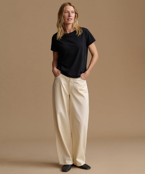 A woman stands against a plain beige studio background wearing a black short-sleeve crew-neck T-shirt and cream wide-leg trousers, with her hands in the trouser pockets.