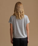 A woman viewed from behind wearing a light gray short-sleeve t-shirt and black pants, standing against a solid tan studio background.