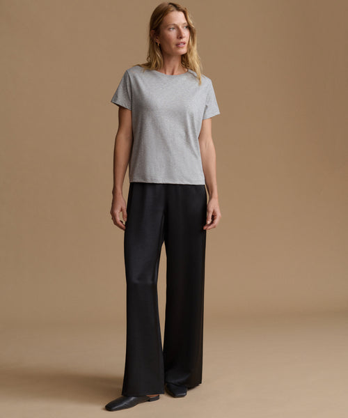 A woman modeling a light gray short-sleeve crewneck t-shirt and black wide-leg trousers against a solid beige studio background.