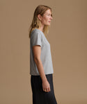 A woman in side profile wearing a light gray short-sleeve knit top and dark navy pants, standing against a solid warm beige studio background.