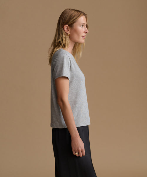 A woman in side profile wearing a light gray short-sleeve knit top and dark navy pants, standing against a solid warm beige studio background.