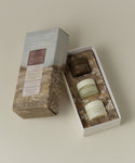 A boxed set of three Oak Essentials skincare products displayed on a warm beige background. The set includes a tall outer box with a floral field illustration and a white inner tray holding two cream-colored jars and one dark brown jar, all labeled Oak Essentials.