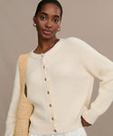 A woman models the Cotton Hayes Crewneck Cardigan in cream, styled with gold hoop earrings, and holds a woven tote bag over her shoulder against a beige background.