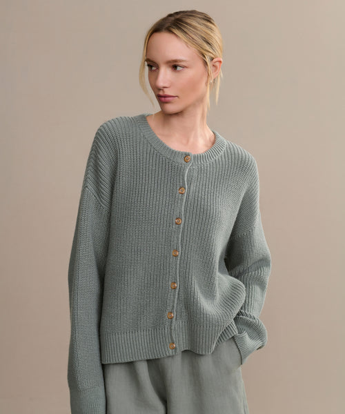 A woman with blonde hair in a low ponytail wears the Cotton Hayes Crewneck Cardigan in sage green with wooden buttons and matching pants, standing against a plain beige background.