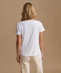 A woman viewed from behind wearing a plain white short-sleeve t-shirt and light beige pants, standing against a solid warm beige studio background.