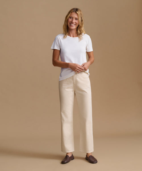 A woman modeling a white short-sleeve crewneck t-shirt and cream wide-leg trousers, paired with dark brown loafers, against a solid beige studio background.