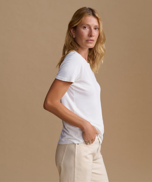 A woman wearing a white short-sleeve crewneck t-shirt and cream-colored trousers, standing against a solid beige studio background.