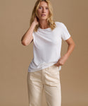 A woman modeling a white short-sleeve crewneck t-shirt paired with cream-colored pants, set against a solid warm beige studio background.