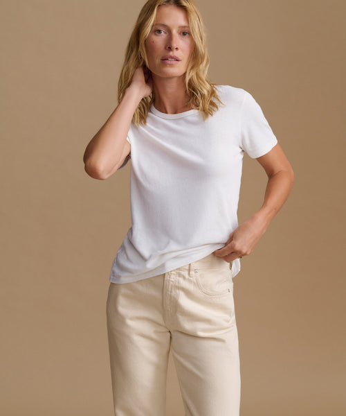 A woman modeling a white short-sleeve crewneck t-shirt paired with cream-colored pants, set against a solid warm beige studio background.