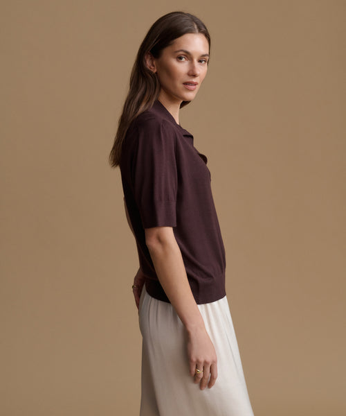 A woman modeling a short-sleeve polo shirt in deep plum purple, paired with a light cream skirt, set against a solid warm beige studio background.