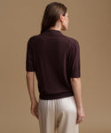 A woman shown from the back wearing a short-sleeve knit polo shirt in deep plum purple and cream trousers, set against a solid warm beige studio background.