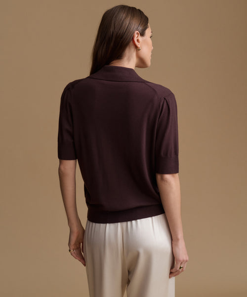 A woman shown from the back wearing a short-sleeve knit polo shirt in deep plum purple and cream trousers, set against a solid warm beige studio background.