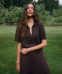 A woman standing outdoors on a grassy lawn wearing a dark brown short-sleeve collared top and matching pleated skirt, with trees in the background.