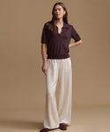 A woman modeling a short-sleeve polo shirt in deep plum brown paired with wide-leg cream trousers, standing against a solid beige studio background.