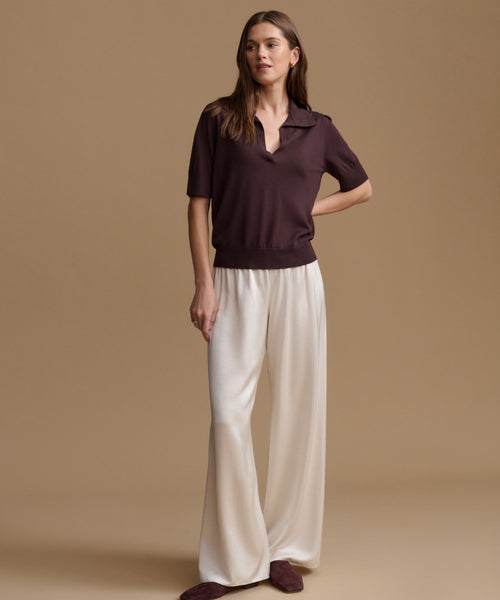 A woman modeling a short-sleeve polo shirt in deep plum brown paired with wide-leg cream trousers, standing against a solid beige studio background.