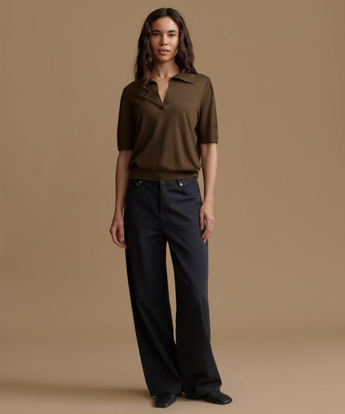A woman stands against a beige studio background wearing a short-sleeve dark olive-brown polo shirt and dark charcoal wide-leg trousers.