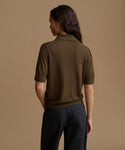 A woman shown from the back wearing a short-sleeve knit polo shirt in olive brown and dark trousers, set against a solid warm beige studio background.
