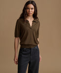 A woman modeling a short-sleeve knit polo shirt in dark brown, paired with dark charcoal pants, against a solid warm beige studio background.