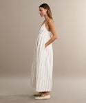 A woman with straight, light brown hair wears the Cove Dress—a long, sleeveless dress with thin gold stripes and delicate shoulder straps—paired with cream flats, against a beige background that highlights the dress’s effortless style.